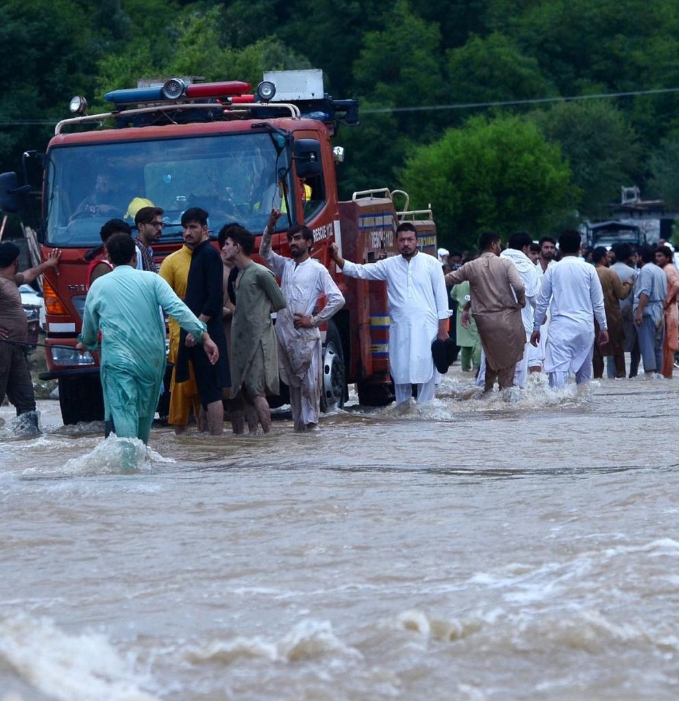 Menschen in Pakistan stehen auf einer überfluteten Straße bei einem Rettungsfahrzeug. Foto: Hussain Ali