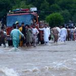 Menschen in Pakistan stehen auf einer überfluteten Straße bei einem Rettungsfahrzeug. Foto: Hussain Ali