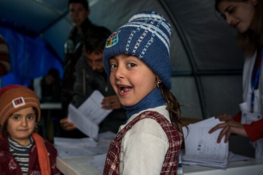 Irak: Im Vordergrund stehen zwei Kinder in warmen Klamotten, das vordere lacht. Im Hintergrund blättern Mitarbeitende in Papieren. Im Irak leistet Ärzte der Welt medizinische Hilfe. © Guillaume Pinon