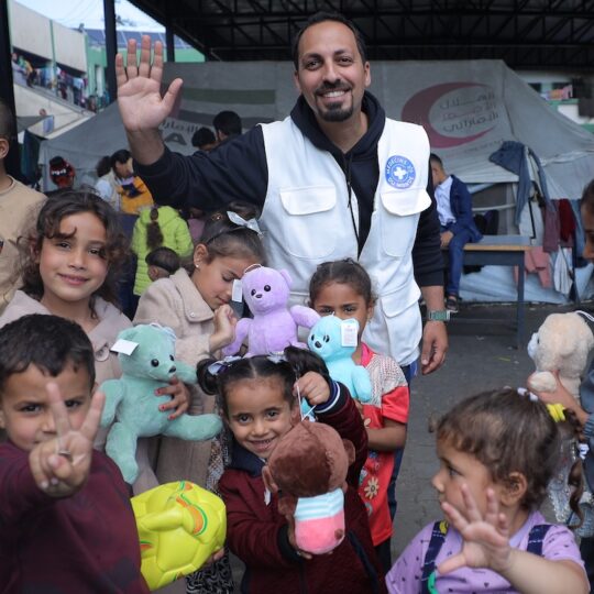 Gaza: Ein Mitarbeiter von Ärzte der Welt winkt in die Kamera. Er ist umgeben von Kindern mit neuen Kuscheltieren.