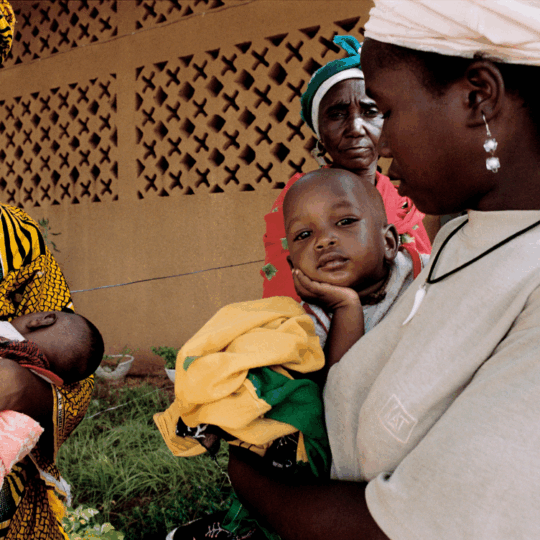 Zwei Frauen tragen ihre Kinder auf dem Arm. Der eine kleine Junge stützt seinen Kopf auf seiner Hand ab und schaut in die Kamera. ©Isabelle Eshragi