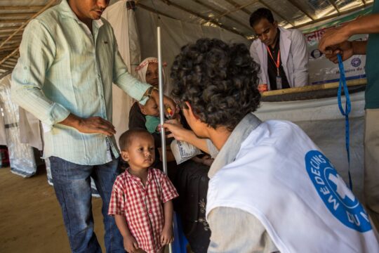 Bangladesch: Ein Mann steht hinter seinem kleinen Sohn in einem Gesundheitszentrum in Bangladesch. Ein Mitarbeiter von Ärzte der Welt hockt vor dem Jungen und misst seine Größe. © Ärzte der Welt