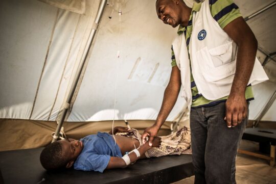 Haiti: Ein Mitarbeiter von Ärzte der Welt hält die Hand eines Jungen, der auf einer Liege liegt und eine Infusion hat. © Olivier Papegnies