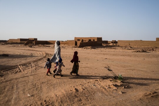Niger: Eine Mutter mit zwei Kinder läuft durch eine sandige Landschaft nahe Agadez. © Kristof Vadino