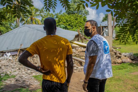 Ein Haitianer und ein Mitarbeiter von Ärzte der Welt unterhalten sich, während sie vor einem zerstörten Gebäude stehen. © Berthony Raymond