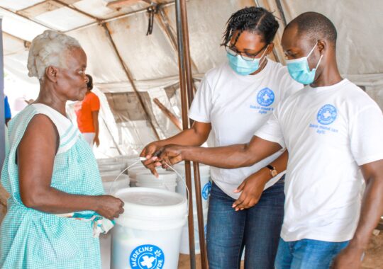 Ein Mitarbeiter und eine Mitarbeiterin von Ärzte der Welt überreichen eine Haitianerin einen Eimer mit Hygieneartikeln. © Berthony Raymond