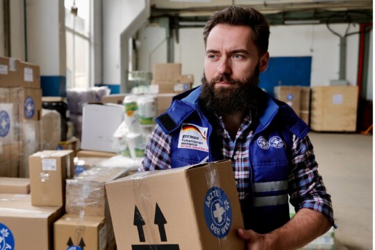 Ukraine: Ein Mitarbeiter von hält ein Paket mit Hilfsgütern in der Hand. Hinter ihm türmen sich noch viele weitere Pakete in einer Lagerhalle. © Till Mayer