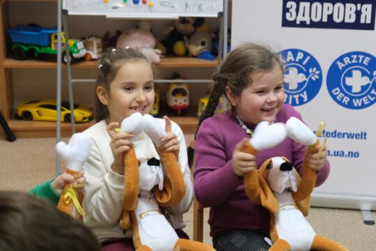Zwei Mädchen bei der Hibuki-Therapie in der Ukraine: Sie halten ihre Stofftiere in der Hand und lächeln. © Ärzte der Welt