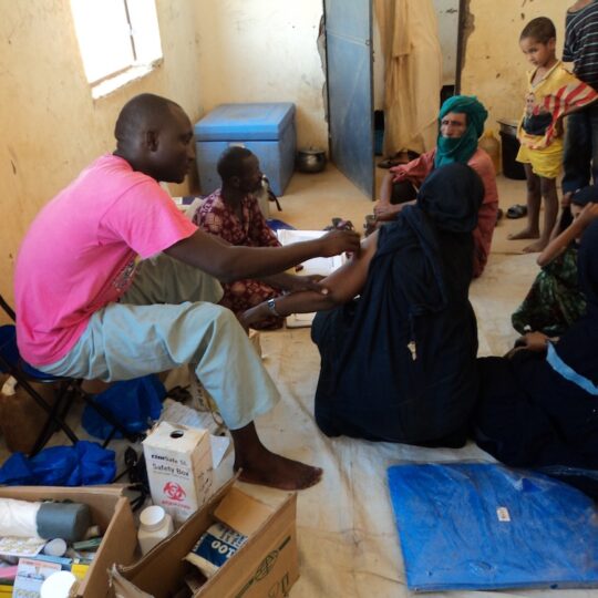 Ein Raum voller Patient*innen und Kinder in einer medizinischen Einrichtung von Ärzte der Welt. Ein Mitarbeiter impft eine Frau, die ihren Arm ausstreckt.