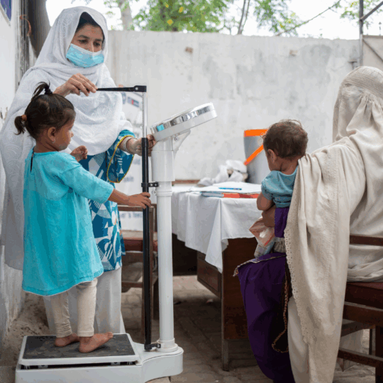 Pakistan: Eine Mitarbeiterin von Ärzte der Welt misst die Größe eines kleinen Mädchens. Die Mutter sitzt auf einem Stuhl daneben und hält ihr anderes Kind auf dem Arm. ©Saiyna Bashir