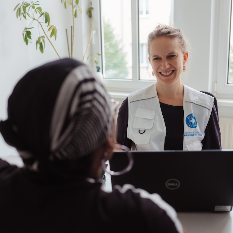 Sprechstundenzimmer in einer Arztpraxis, eine Frau mit Weste von Ärzte der Welt lächelt über den Schreibtisch.