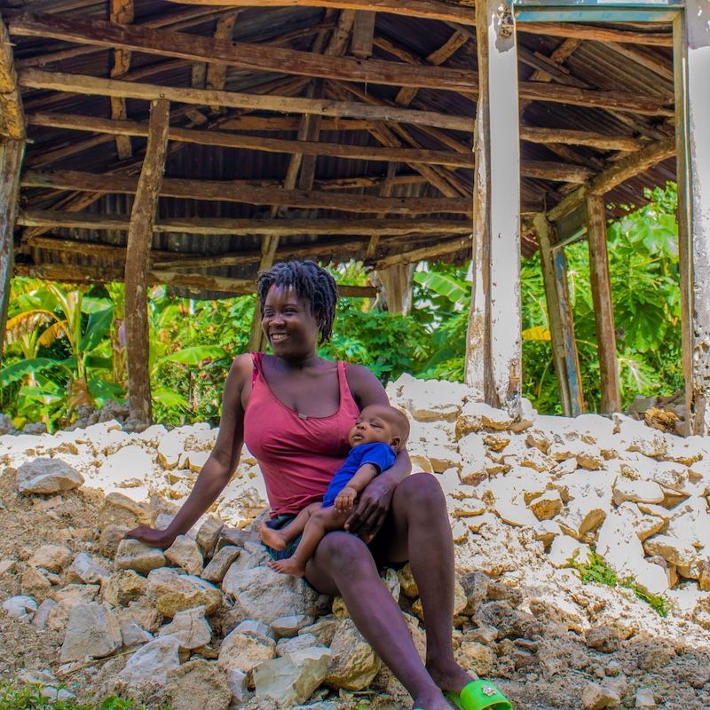 Eine Mutter und ihr Baby sitzen vor einer Hütte. © Berthony Raymond