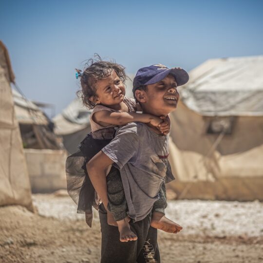 Syrien: Junge trägt kleines Mädchen auf dem Rücken, in einem Zeltlager. Ärzte der Welt leistet medizinische Hilfe in Syrien.