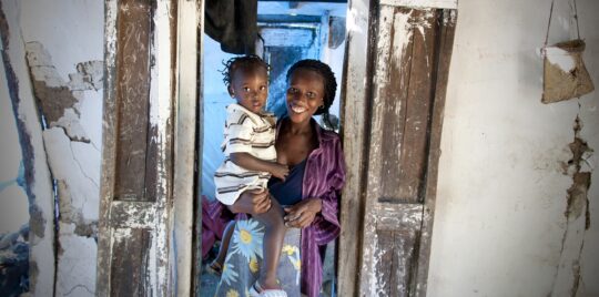 © Pierre-W. Henry Eine Frau steht in einem Türeingang, auf ihrem Arm ein Kleinkind. Ärzte der Welt leistet medizinische Hilfe in Haiti.