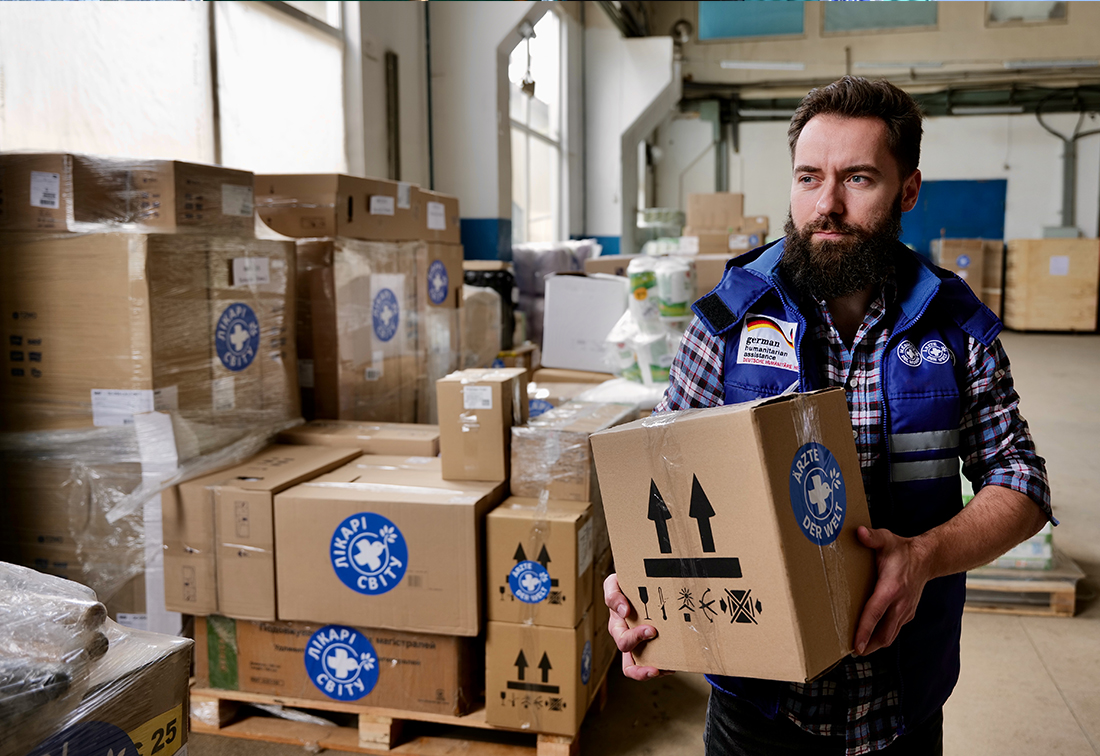 Ukraine: Ein Mitarbeiter von hält ein Paket mit Hilfsgütern in der Hand. Hinter ihm türmen sich noch viele weitere Pakete in einer Lagerhalle. © Till Maye