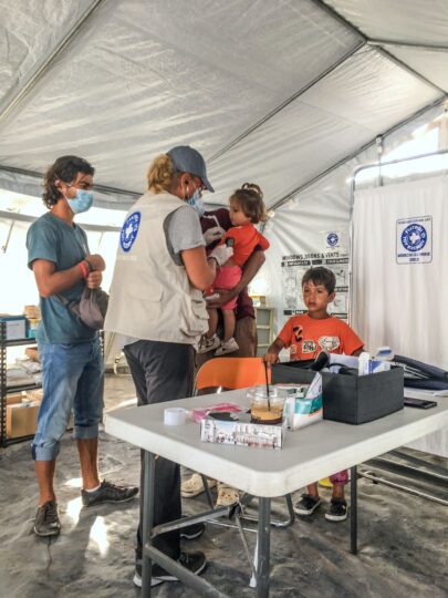 Zwei Männer stehen mit ihren Kindern in einem Behandlungszelt von Ärzte der Welt. Eine Ärztin impft das kleine Mädchen. Ärzte der Welt leistet in Griechenland medizinische Notfallhilfe. © Ärzte der Welt