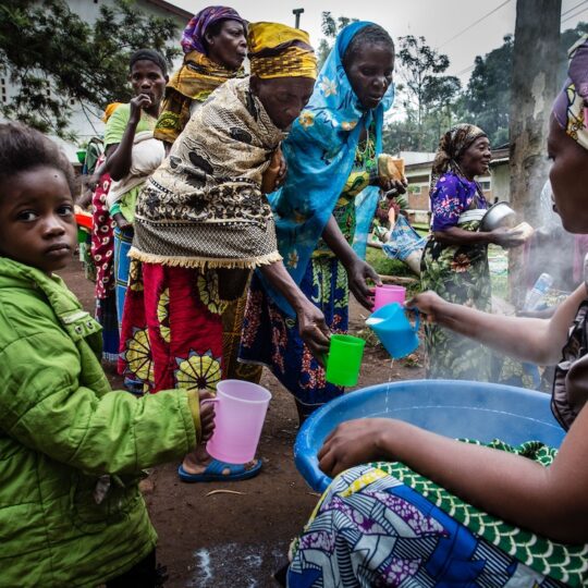 Kongo: Eine Frau schöpft heiße Milch für andere Frauen und ein kleines Mädchen aus.