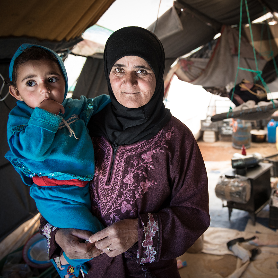 Eine Frau im Kopftuch hält ein Kind auf dem Arm. Sie stehen in einer kargen Hütte.