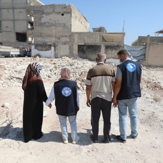 Ein Mitarbeiter und eine Mitarbeiterin von Ärzte der Welt stehen einer Frau und einem Mann bei, während sie vor einem zerstörten Gebäude in Syrien stehen.