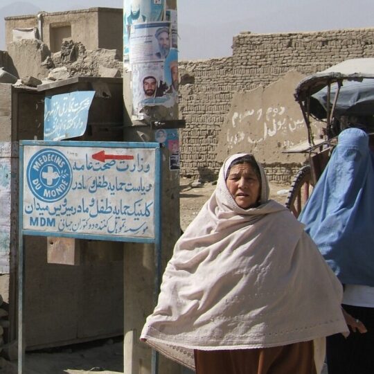 Zwei Frauen stehen vor einem zerstörten Gebäude in Afghanistan. Hinter ihnen weist ein Schild den Weg zur medizinischen Einrichtung von Ärzte der Welt. © Isabelle Eshraghi