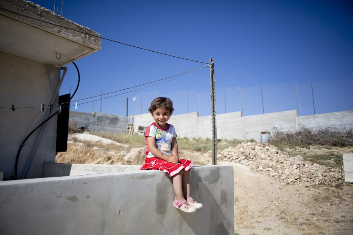 Les enfants d'Asmahan Yaya Safdi jouent dans leur jardin devant le mur construit pour les proteger des colons de Yizhar. Palästinensische Gebiete: Ein kleines Mädchen sitzt auf einer Mauer und lacht, im Hintergrund sieht man Mauern und Stacheldraht, der Himmel ist strahlend blau. © Bruno Fert