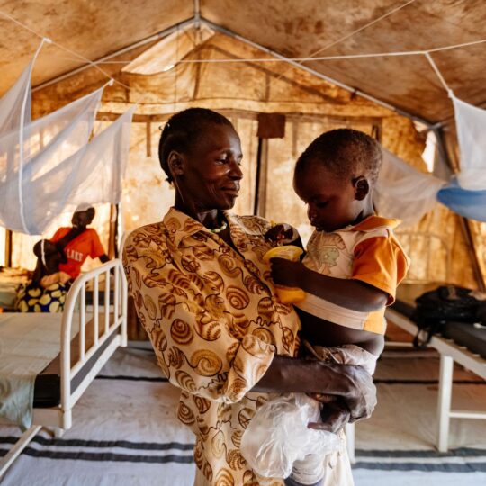 Uganda: Eine Frau mit einem Kleinkind auf dem Arm, in einer Notunterkunft mit Betten. Ärzte der Welt leistet medizinische Hilfe in Uganda. © Sébastien Duijndam
