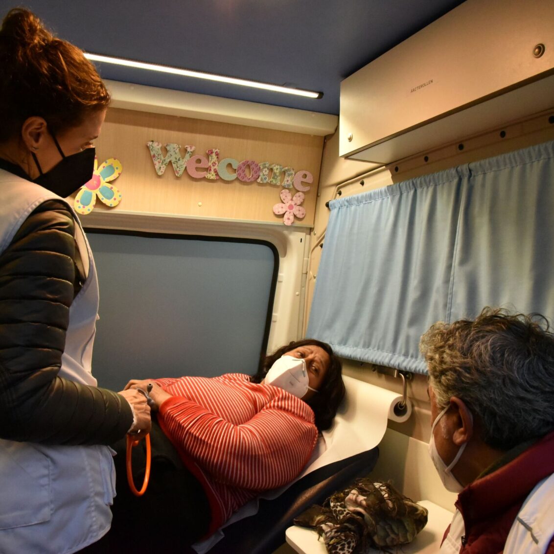 Eine Patientin liegt im Münchner Behandlungsbus von Ärzte der Welt auf einer Behandlungsliege. Zwei Mitarbeiter*innen sprechen mit ihr. © Chris Schmid
