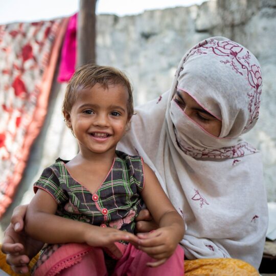 Pakistan: Ein lächelndes Kleinkind sitzt auf dem Schoß einer Frau. Ärzte der Welt leistet medizinische Hilfe in Pakistan, unter anderem für Mütter und Kinder. © Saiyan Bashir