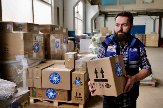 Ukraine: Ein Mitarbeiter von hält ein Paket mit Hilfsgütern in der Hand. Hinter ihm türmen sich noch viele weitere Pakete in einer Lagerhalle. © Till Mayer