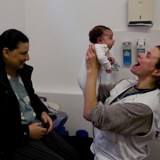 München: Eine Ärztin hebt ein Baby hoch und lacht es an, die Mutter sitzt daneben und lächelt. Ärzte der Welt bietet in Deutschland Kindersprechstunden. © Till Mayer