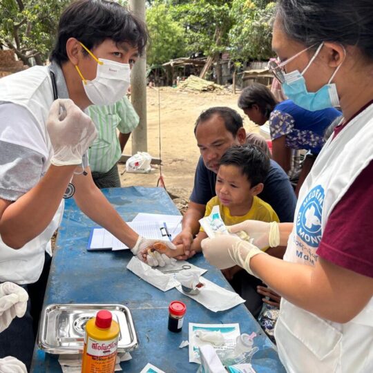 Nach dem Erdbeben in Myanmar: Mitarbeiter*innen von Ärzte der Welt versorgen die verletzte Hand eines kleinen Jungen, der auf dem Schoß seines Vaters sitzt. © Ärzte der Welt