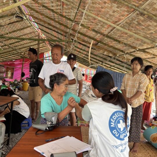 Eine mobile Klinik in Myanmar: Eine Mitarbeiterin von Ärzte der Welt spricht mit einer Patientin. Um sie herum stehen weitere Einwohner*innen. © Ärzte der Welt
