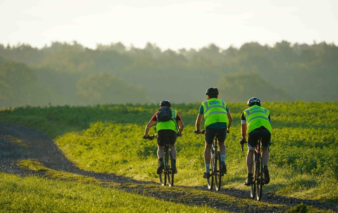 Drei Teilnehmende der Fahrrad-Spendenaktion Doc’Riders für Ärzte der Welt in Belgien. Sie tragen gelbe Westen und fahren über eine grüne Wiese. © Ärzte der Welt