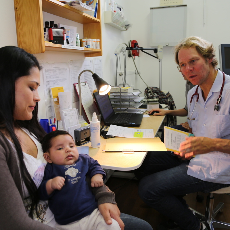 Kindersprechstunde in München: Ein Arzt unterhält sich mit einer Frau, sie hat ein Baby auf dem Arm. © Mike Yousaf
