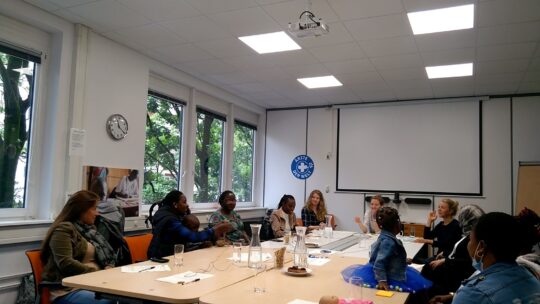 Workshop des reach.out Projekts in Deutschland. Mehrere Frauen und Kinder sitzen um einen großen Tisch und unterhalten sich fröhlich. © Ärzte der Welt