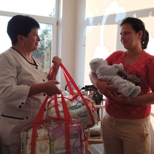 Eine Mitarbeiterin von Ärzte der Welt überreicht einer ukrainischen Mutter mit einem Baby im Arm ein Paket mit Ausstattung für Neugeborene. © Ärzte der Welt