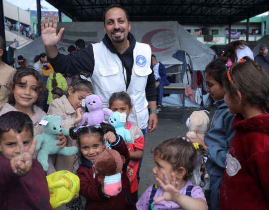 Palästina/Gaza: Ein Mitarbeiter von Ärzte der Welt steht mit einer Gruppe Kinder zusammen. Die Kinder halten Stofftiere in der Hand und lächeln in die Kamera. © Ärzte der Welt