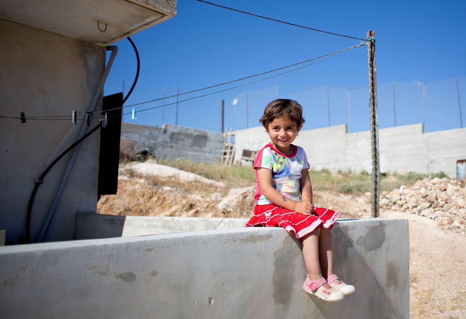 Palästinensische Gebiete: Ein kleines Mädchen sitzt auf einer Mauer und lacht, im Hintergrund sieht man Mauern und Stacheldraht, der Himmel ist strahlend blau. © Bruno Fert