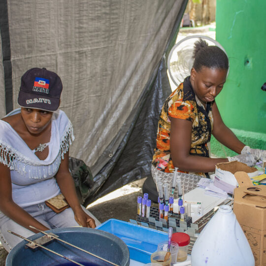 Haiti: Zwei Frauen sitzen an einem Tisch mit einem Mikroskop und medizinischer Ausrüstung. © Berthony Raymond