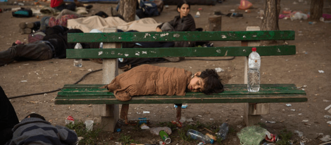 ©Olmo Calvo Foto aus der Balkanroute. Mehrere Personen versuchen sich in einer zugemüllten Gegend auszuruhen. Ein Frau sitzt auf und schaut in die Ferne. Auf der Bank liegt das einzige Kind, was dort zu sehen ist und schläft.