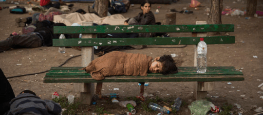 Foto aus der Balkanroute. Mehrere Personen versuchen sich in einer zugemüllten Gegend auszuruhen. Ein Frau sitzt auf und schaut in die Ferne. Auf der Bank liegt das einzige Kind, was dort zu sehen ist und schläft.