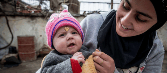 Irak: Glückliche Mutter und ihr Baby in eher winterlicher Kleidung. Baby tragt Kette am Kopf.