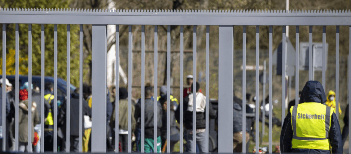 © Heiko Becker, HMB-Media Person in Warnweste steht vor einem soliden Zaun. Hinter dem Zaun befinden sich Geflüchtete