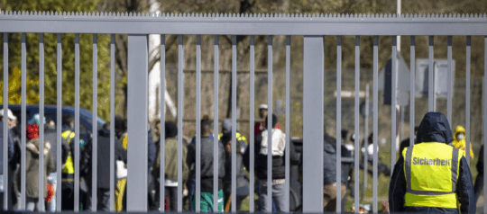 Person in Warnweste steht vor einem soliden Zaun. Hinter dem Zaun befinden sich Geflüchtete