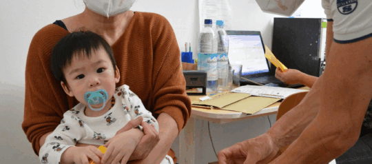 Ein sehr junges Kind und dessen Mutter befinden sich in einer medizinischen Sprechstunde mit open.med. Das Kind sitzt auf dem Schoß der Mutter und schaut in die Kamera