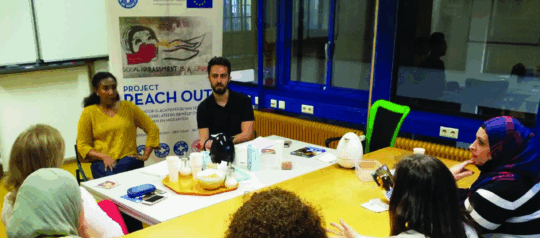 Mehrere Personen sind bei einem reach.out treffen. Im Hintergrund ist ein Banner mit der Aufschrift 