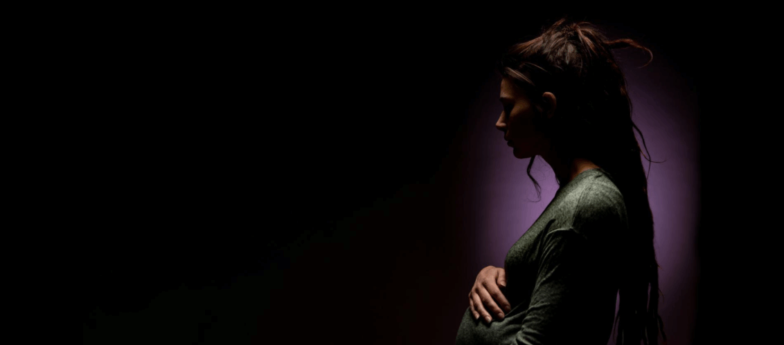 ©COMM-WORK / Eric Langerbeins Schwangere Frau innerhalb einer dunklen Leere schaut leicht besorgt und fässt sich an den Bauch.