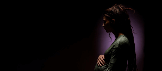 Schwangere Frau innerhalb einer dunklen Leere schaut leicht besorgt und fässt sich an den Bauch.