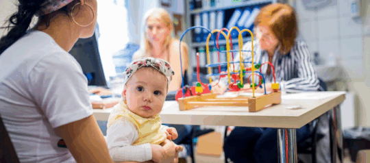 Mehrere Personen befinden sich in einer open.med Kindersprechstunde. Ein Baby, welches auf dem Schoß seiner Mutter sitzt, schaut direkt in die Kamera